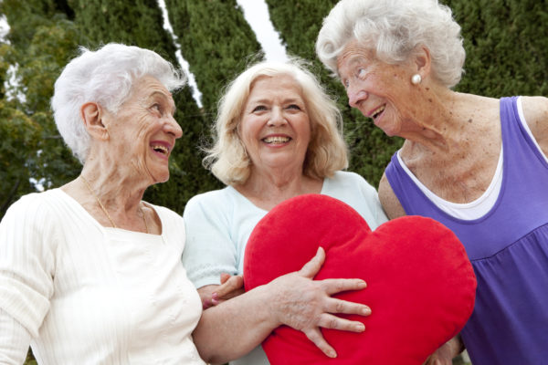 three senior woman standing together; middle one holds a red heart pillow