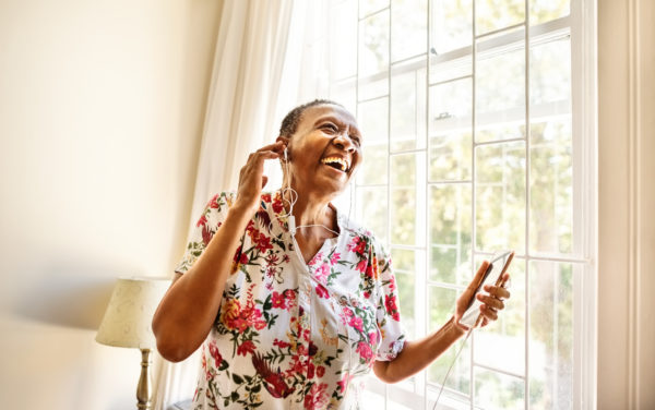 Cheerful senior female standing by window enjoying listening to music on earphones