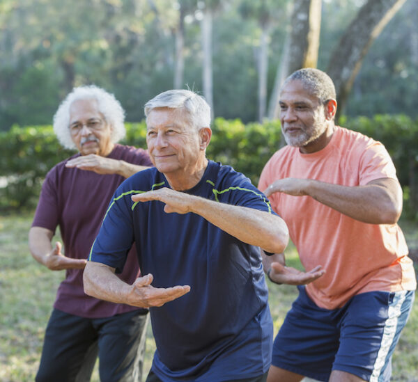 A group of three multi-ethnic senior men in the park practicing tai chi. The focus is on the caucasian man standing in the middle. He is in his 70s.